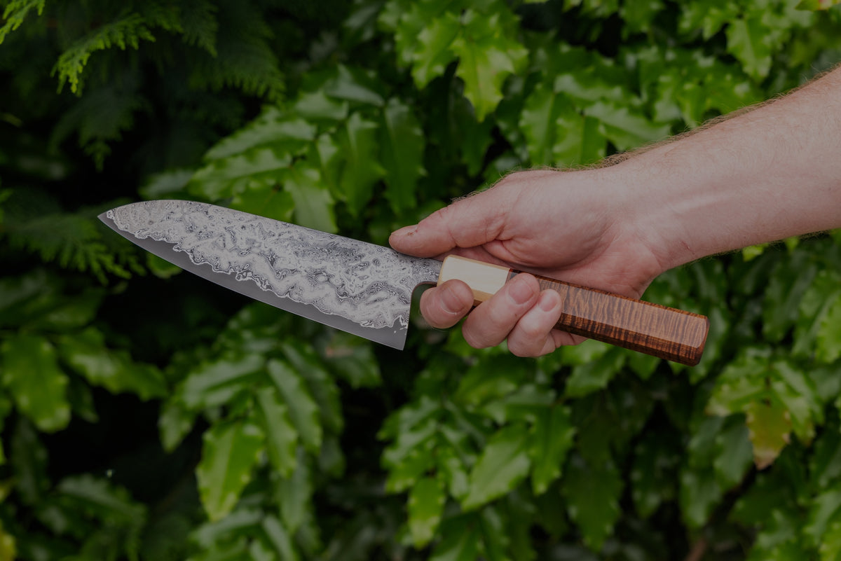 A hand holding a Damascus chef knife in front of green leaves